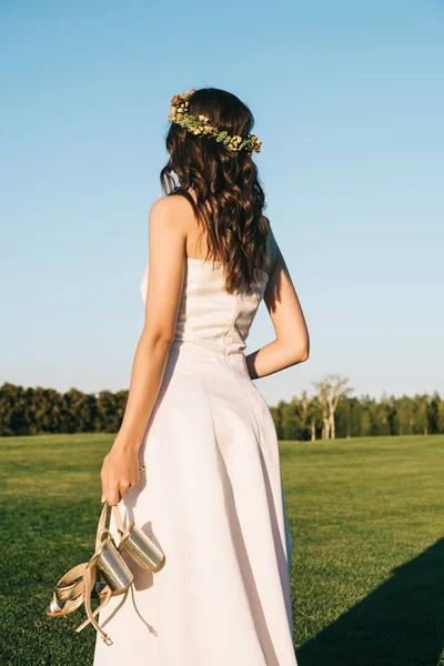 Вид сзади на красивую нежную молодую невесту, держащую туфли на зеленой ленточке в парке — стоковое фото