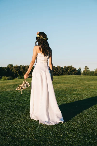Вид ззаду ніжної молодої нареченої, що тримає взуття і ходить на зеленому газоні — стокове фото