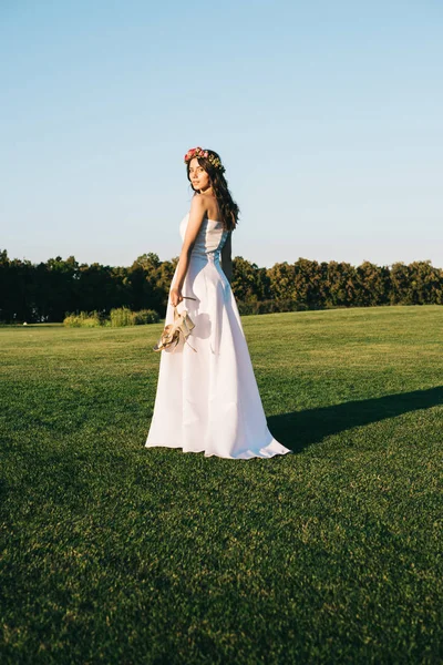 Bela jovem noiva segurando sapatos e olhando para a câmera enquanto está em pé no gramado verde — Fotografia de Stock
