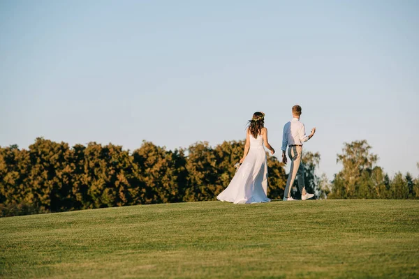 Visão traseira do belo casal jovem casamento andando no prado verde no parque — Fotografia de Stock