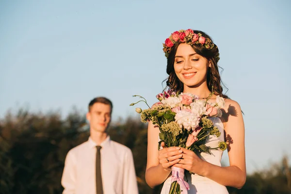 Hermosa feliz joven novia celebración de ramo de boda y novio de pie detrás - foto de stock