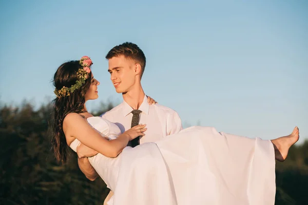 Happy young groom carrying beautiful bride against blue sky — Stock Photo