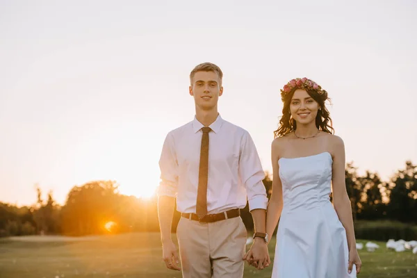 Красива щаслива молода весільна пара тримає руки і посміхається на камеру під час прогулянки в парку на заході сонця — стокове фото