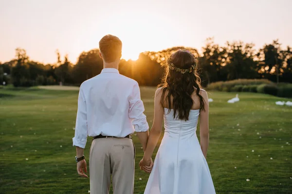 Visão traseira do jovem casal de casamento de mãos dadas e de pé juntos no parque ao pôr do sol — Fotografia de Stock