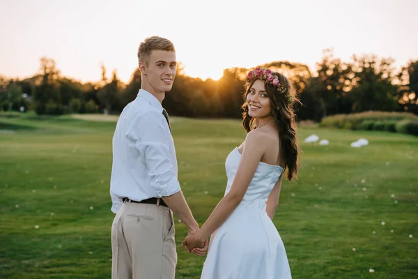 Красивая счастливая молодая свадебная пара, держащаяся за руки и улыбающаяся перед камерой в парке на закате — стоковое фото