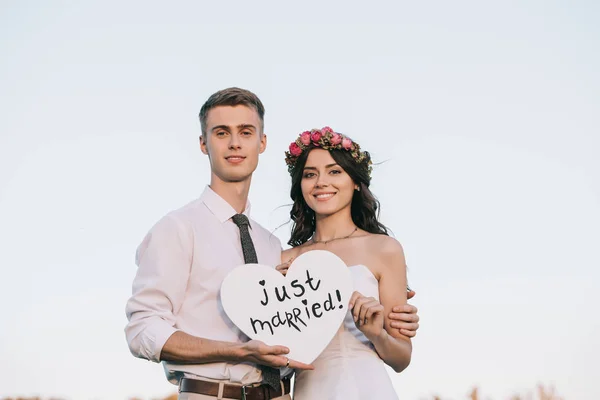 Happy young wedding couple holding heart with just married inscription and smiling at camera — Stock Photo