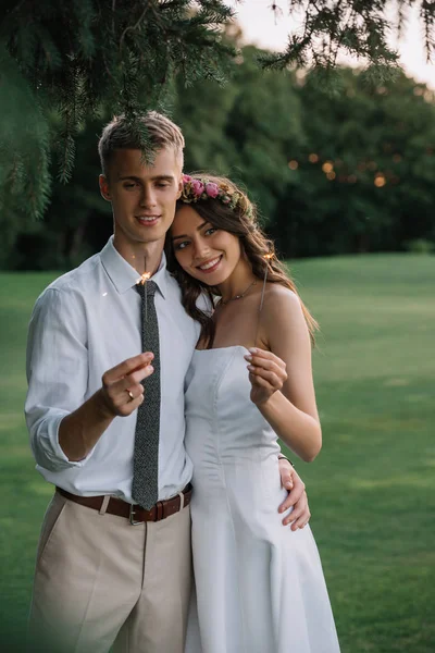 Beau heureux jeune couple de mariage tenant des étincelles et souriant à l'extérieur — Photo de stock