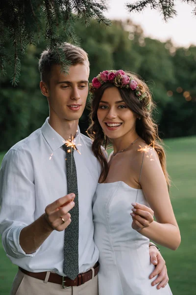 Beautiful romantic young wedding couple holding sparklers and smiling at camera — Stock Photo