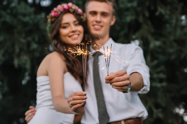 Vista de cerca de feliz joven pareja de boda sosteniendo chispas y sonriendo a la cámara - foto de stock