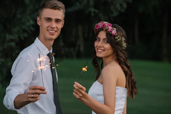 Beautiful happy young wedding couple holding sparklers and smiling at camera — Stock Photo