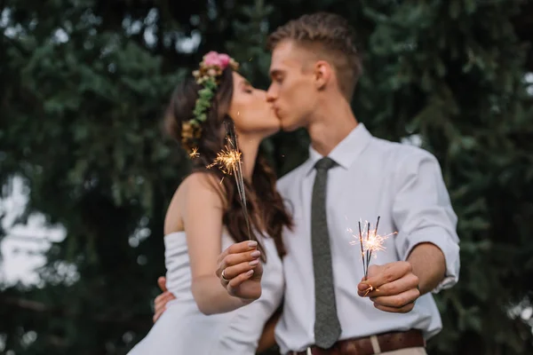 Избирательный фокус счастливой молодой свадебной пары, держащей искры и целующейся — стоковое фото