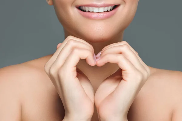 Foto recortada de una mujer sonriente con dientes blancos mostrando el signo del corazón con las manos aisladas en gris - foto de stock