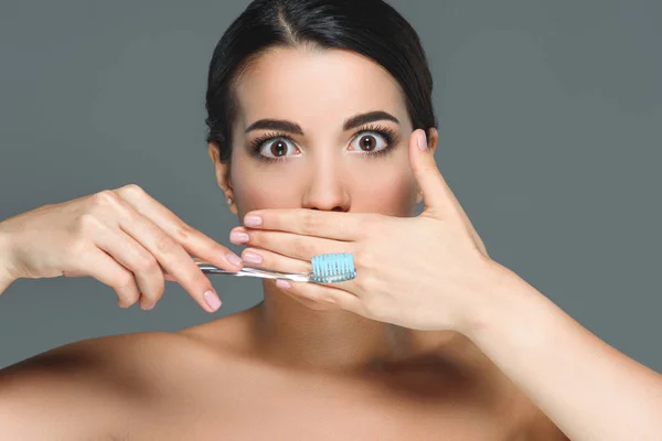 Brunette woman with bare shoulders and tooth brush covering mouth isolated on grey — Stock Photo