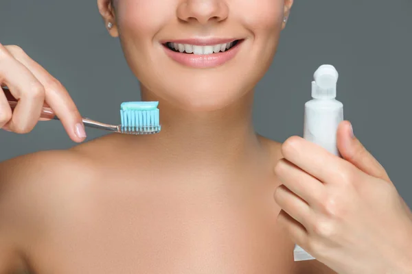 Partial view of smiling woman with tooth brush and tooth paste tube in hands isolated on grey — Stock Photo