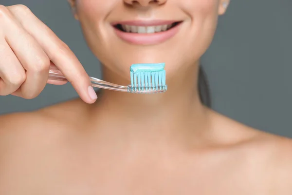 Частковий вид усміхненої жінки, що тримає зубну щітку з зубною пастою ізольовано на сірому — стокове фото