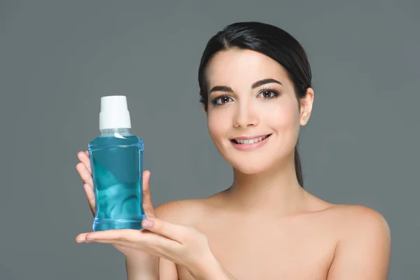 Portrait of smiling woman with bare shoulders holding mouthwash isolated on grey — Stock Photo