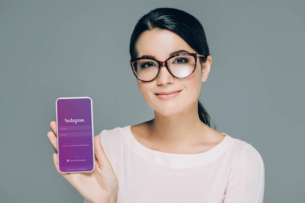 Портрет усміхається жінка в окулярів показ смартфон з instagram app на екрані ізольовані на сірому — стокове фото