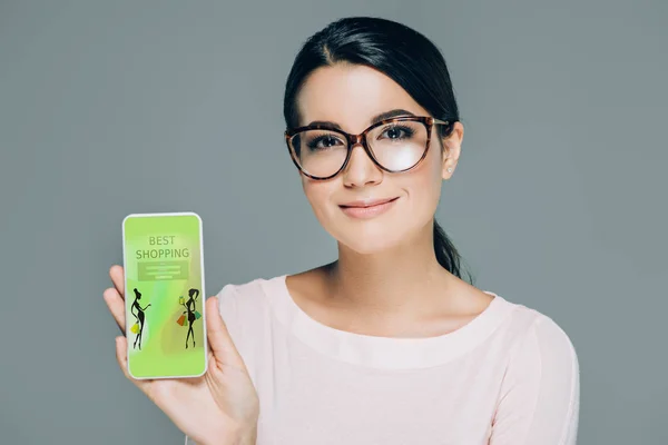Porträt einer lächelnden Frau mit Brille, die Smartphone mit bester Shopping-Website auf dem Bildschirm zeigt, isoliert auf grau — Stockfoto