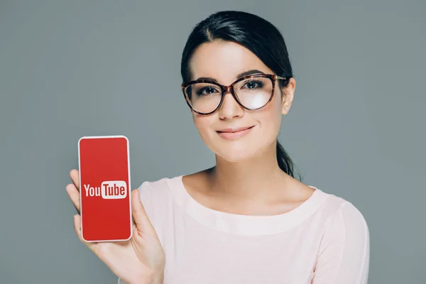 Портрет усміхається жінка в окулярів показ смартфон з сайту youtube екран ізольовані на сірому — стокове фото