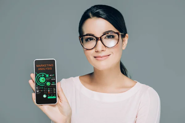 Портрет улыбающейся женщины в очках, показывающий смартфон с маркетинговым анализом на экране, изолированном на сером — стоковое фото