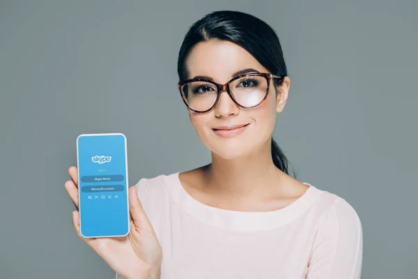 Portrait of smiling woman in eyeglasses showing smartphone with skype app on screen isolated on grey — Stock Photo