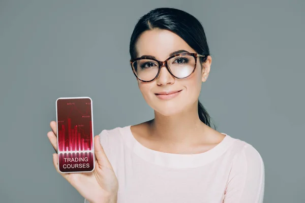 Портрет усміхненої жінки в окулярах, що показує смартфон з логотипом торгових курсів на екрані ізольовано на сірому — стокове фото