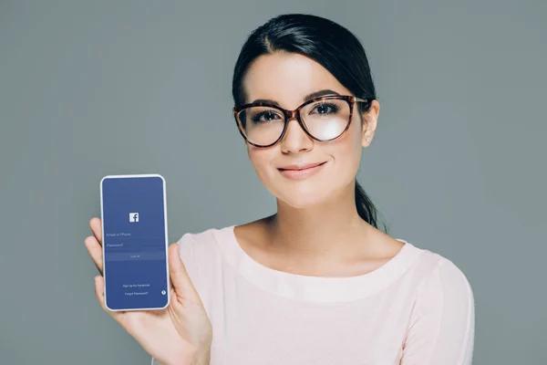 Портрет усміхається жінка в окулярів показ смартфон з facebook логотип на екрані ізольовані на сірому — стокове фото