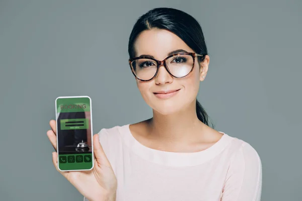 Retrato de mujer sonriente en gafas que muestra el teléfono inteligente con la página web de reserva en la pantalla aislada en gris - foto de stock