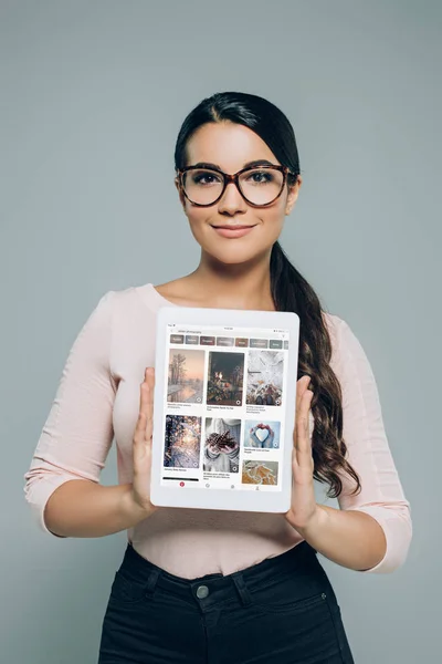 Retrato de mujer morena sonriente en anteojos mostrando tableta con página web pinterest en pantalla en manos aisladas en gris - foto de stock