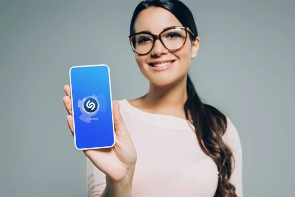 Chica sonriente mostrando teléfono inteligente con aplicación shazam, aislado en gris - foto de stock