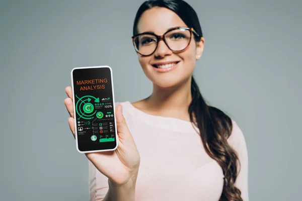 Усміхнена жінка, що показує смартфон з маркетинговим аналізом та інфографікою, ізольована на сірому — стокове фото
