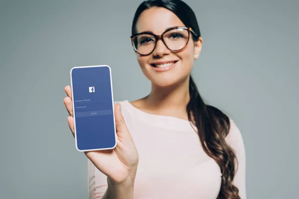 Hermosa chica sonriente presentando teléfono inteligente con aplicación de Facebook, aislado en gris - foto de stock