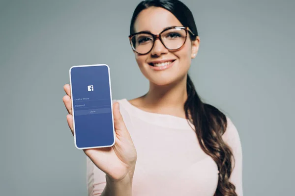 Bella ragazza sorridente che presenta smartphone con app facebook, isolato su grigio — Foto stock