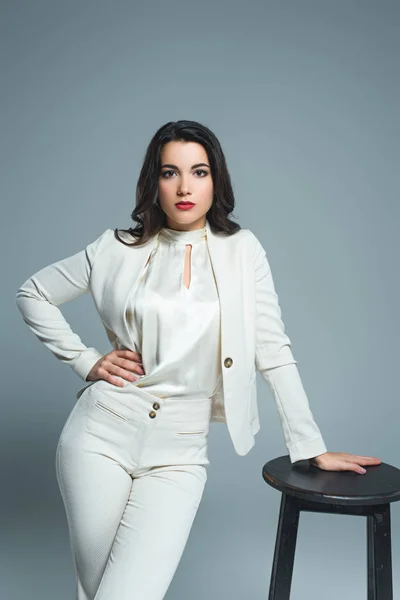 Elegant girl in white suit posing at stool, isolated on grey — Stock Photo