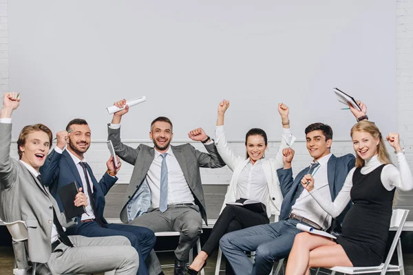 Happy businesspeople with raised hands sitting on chairs at training in hub — Stock Photo