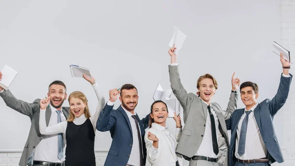 Счастливые бизнесмены, стоящие с поднятыми руками и держащие блокноты в центре — стоковое фото
