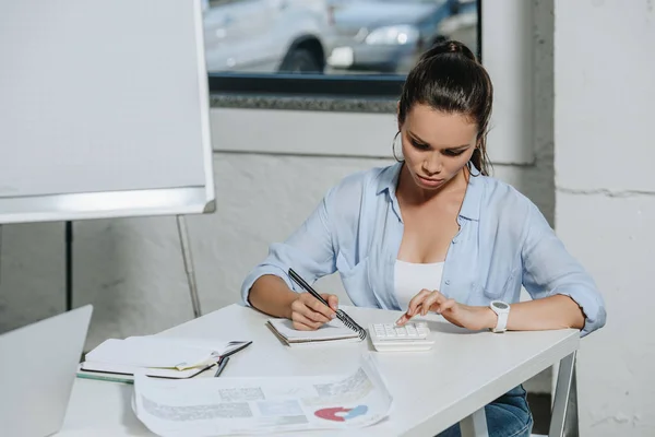 Привлекательная деловая женщина, использующая калькулятор и пишущая что-то в ноутбук в офисе — стоковое фото