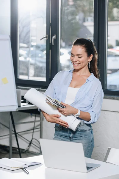 Sonriente atractiva empresaria sosteniendo planos y cuadernos en la oficina - foto de stock