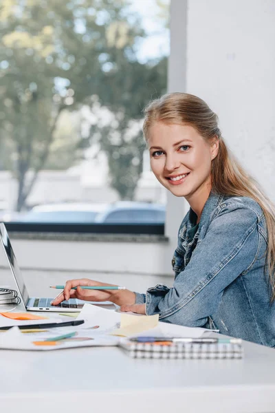 Sonriente mujer de negocios trabajando con el ordenador portátil en la oficina y mirando a la cámara - foto de stock