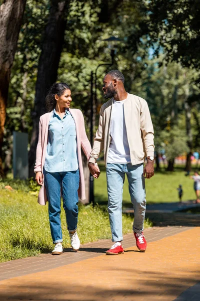 Улыбающаяся африканская американская пара, держащаяся за руки и идущая в парке — стоковое фото