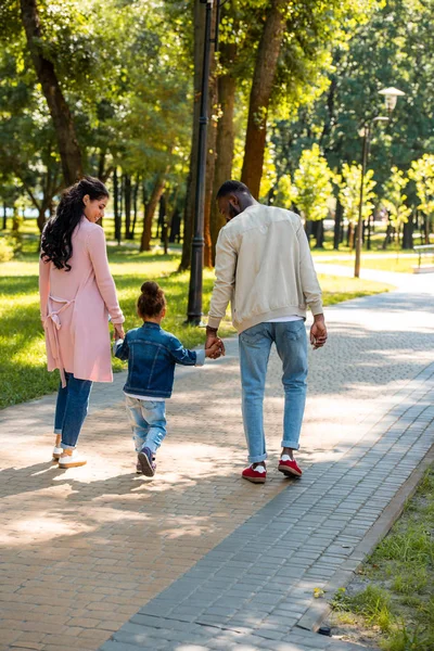 Vista posterior de los padres afroamericanos y su hija tomados de la mano y caminando en el parque - foto de stock