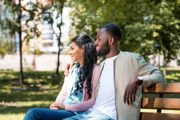 Smiling african american couple sitting on wooden bench in park and looking away — Stock Photo