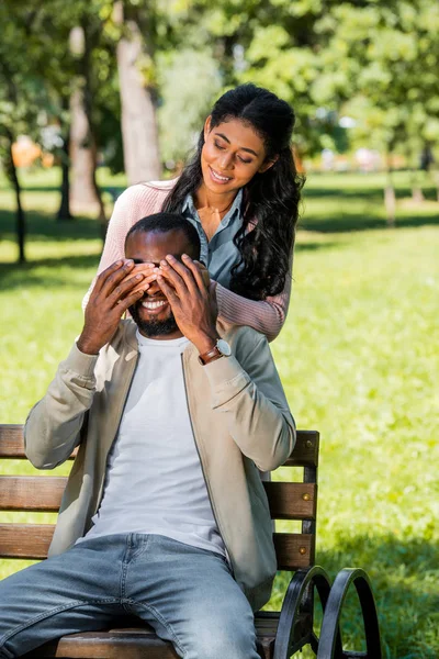 African american girlfriend closing boyfriend eyes while he sitting on bench in park — Stock Photo