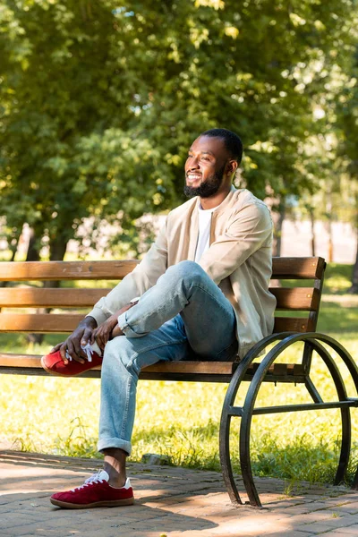 Guapo sonriente afroamericano hombre sentado en banco de madera en el parque y mirando hacia otro lado - foto de stock