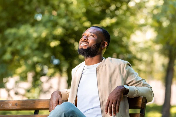 Guapo sonriente afroamericano hombre sentado en banco de madera en el parque y mirando hacia arriba - foto de stock