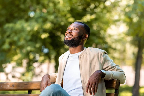 Handsome smiling african american man sitting on wooden bench in park and looking up — Stock Photo