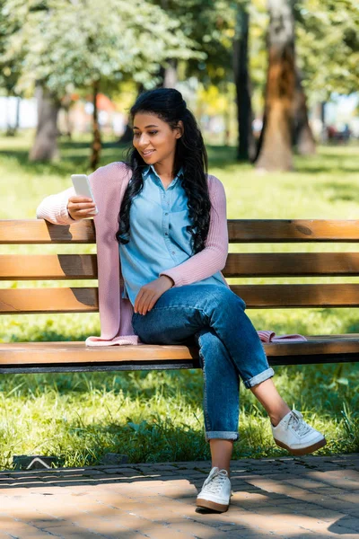 Alegre hermosa mujer afroamericana mirando teléfono inteligente en banco de madera en el parque - foto de stock