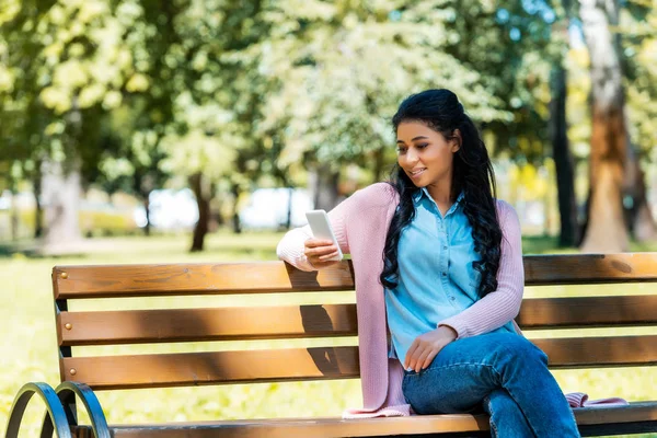 Joyeuse femme afro-américaine attrayante utilisant un smartphone sur un banc en bois dans le parc — Photo de stock