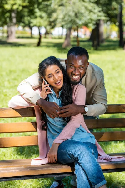 Africano americano novio abrazando novia mientras ella hablando por teléfono inteligente en banco en parque - foto de stock