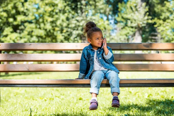 Adorable african american kid sitting on bench in park and talking by smartphone — Stock Photo